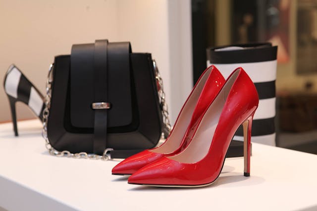 A pair of glossy red high heels displayed on a white surface, accompanied by a black handbag with a silver chain strap and striped accessories in the background.