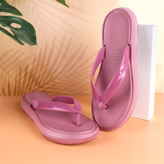 A pair of glossy pink flip-flops displayed against a tropical-themed background. One sandal is propped up on a white block, showing the textured sole, while the other lays flat, highlighting the cushioned footbed and flexible strap.