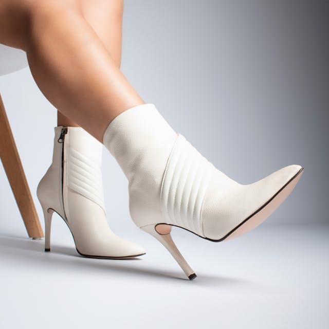 A pair of elegant white ankle boots with pointed toes and slim stiletto heels, worn by a person sitting on a chair. The boots feature a smooth leather finish with ribbed detailing on the upper and a sleek side zipper for easy wear.