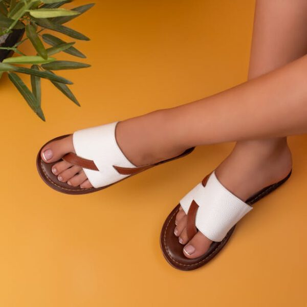 A pair of stylish white and brown slip-on sandals worn by a person sitting on a warm mustard-yellow background. The sandals feature a wide-cut leather strap with a geometric cutout design, a brown sole, and contrast stitching.