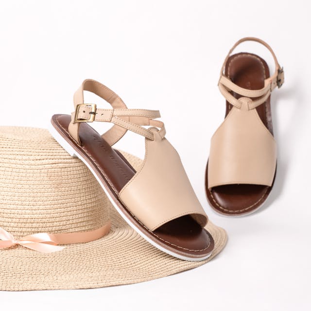 A pair of beige open-toe sandals with an adjustable ankle strap and a cushioned brown footbed, displayed against a white background. One sandal is resting on a straw hat with a beige ribbon, while the other is propped up, showcasing its elegant design.