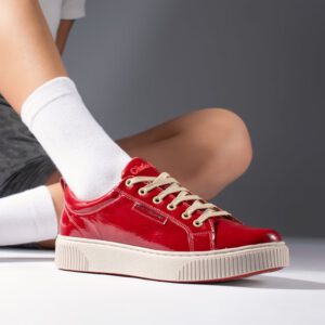 A close-up of a person wearing glossy red low-top sneakers with beige laces and a thick cream-colored rubber sole. The person is sitting with one leg bent, wearing white socks, against a neutral background.