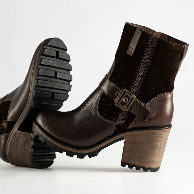 A pair of brown ankle boots with a chunky block heel and rugged rubber sole. The boots feature a mix of smooth leather and suede materials, a decorative side buckle, and a functional zipper closure, set against a clean white background.