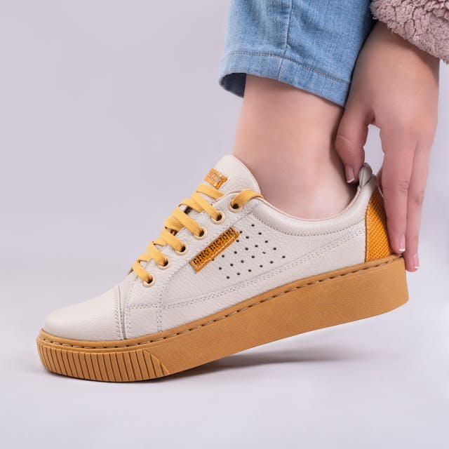 A close-up of a person wearing beige and mustard-yellow sneakers with a textured rubber sole. The shoe has perforated side detailing, mustard-colored laces, and branding accents. The wearer, dressed in light blue jeans, is adjusting the back of the sneaker against a neutral background.
