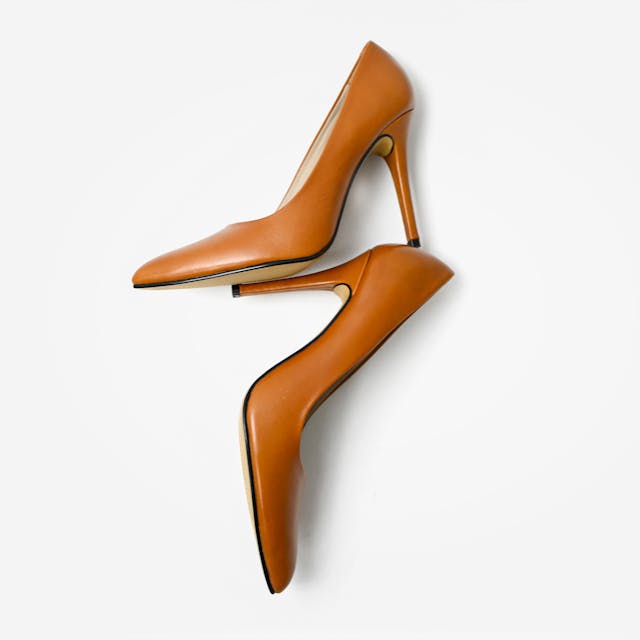 A pair of sleek, tan high-heeled pumps with a pointed toe and stiletto heel, arranged diagonally on a clean white background. The shoes have a smooth, polished finish with a minimalist design.
